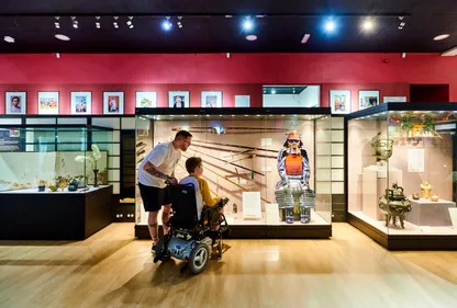 A person looks at a display of Japanese arms and armour with a child who is in a wheelchair..